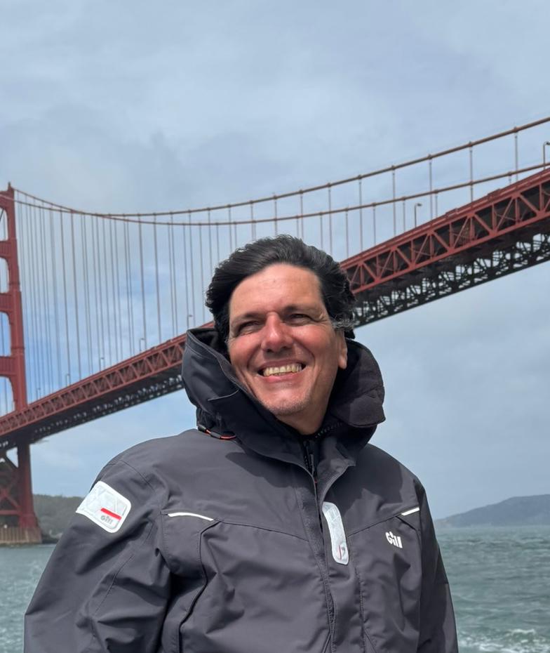Sergio Avellar sorrindo, com roupa de vela escura e a Golden Gate Bridge ao fundo